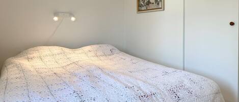 A room with a bed covered in a white knitted blanket, a wall-mounted light fixture, and a framed picture.