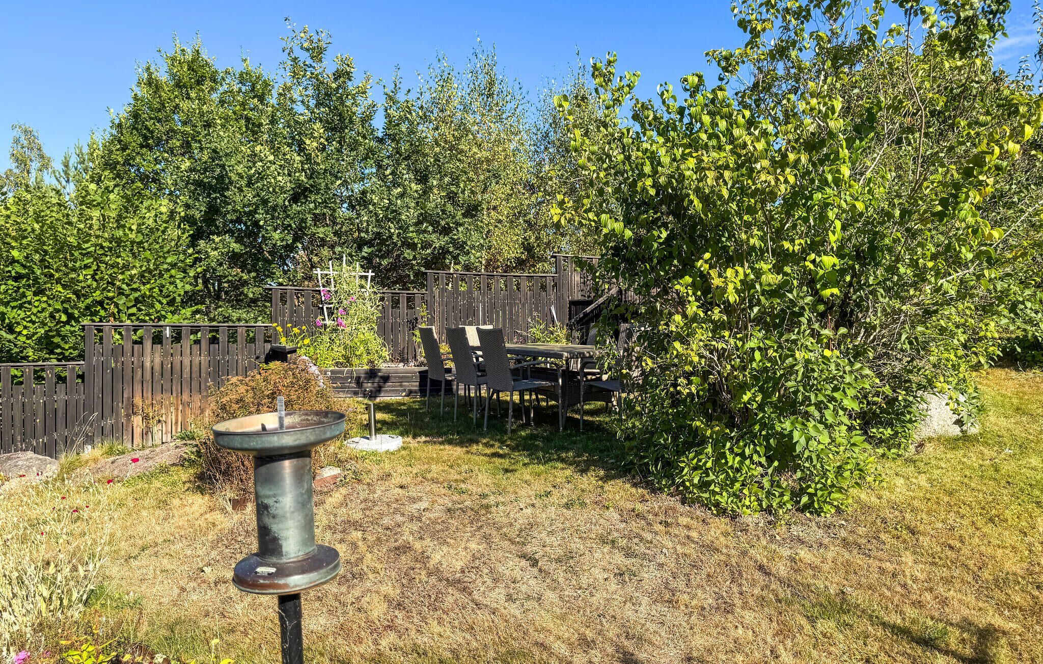 Un jardin avec une clôture en bois, une mangeoire à oiseaux en métal et une table avec des chaises.