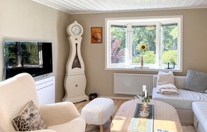 Uma sala de estar com televisão, um relógio branco, uma janela com girassol e um sofá com almofada.