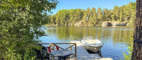 Une jetée en bois, avec un bateau amarré à son extrémité, entourée d’arbres et bordant un lac paisible.