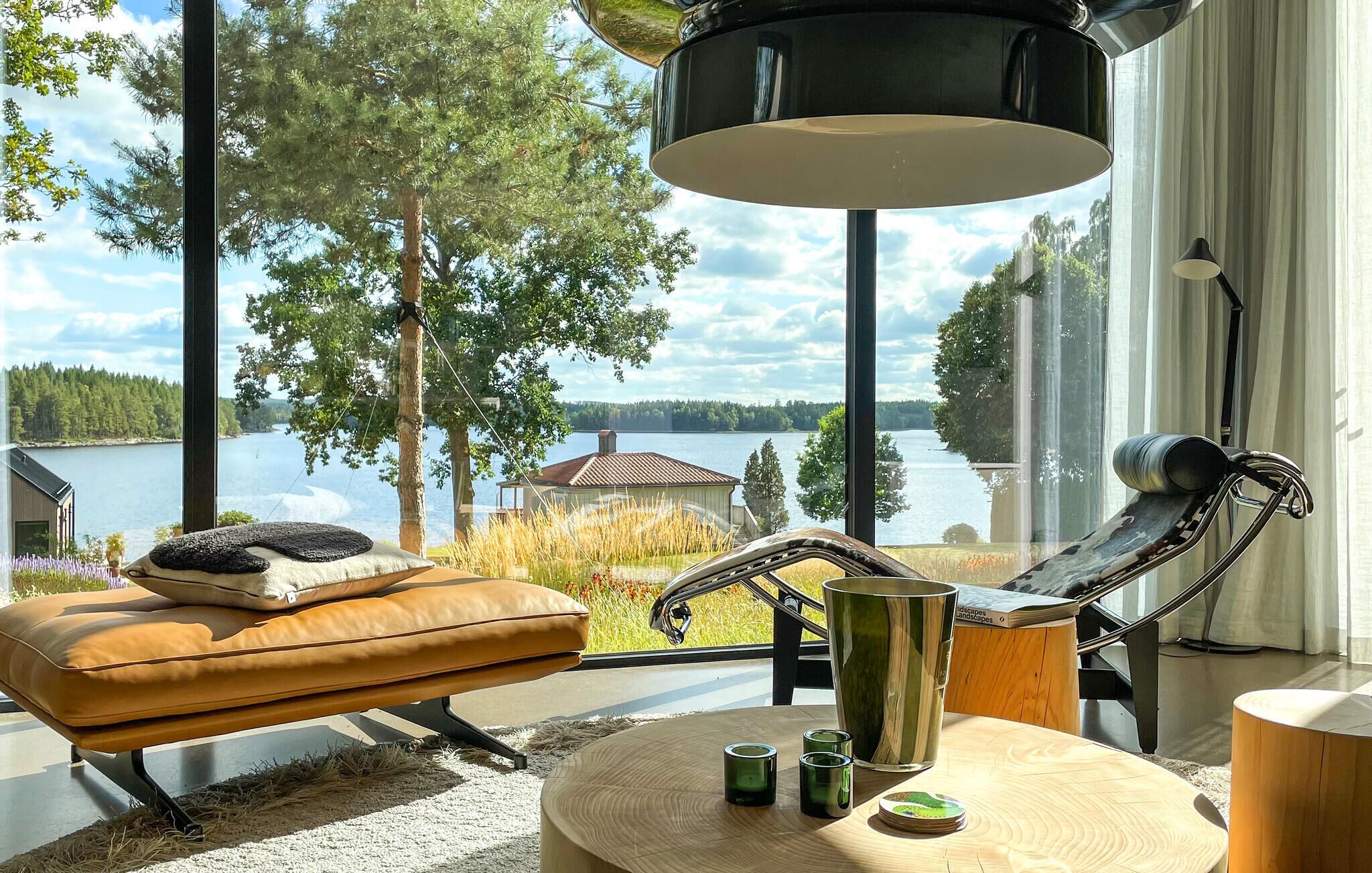A modern living room with a view of a lake, a leather sofa, a metal chair, and a round wooden table with candles.