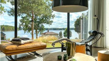 A modern living room with a view of a lake, a leather sofa, a metal chair, and a round wooden table with candles.