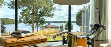 A modern living room with a view of a lake, a leather sofa, a metal chair, and a round wooden table with candles.