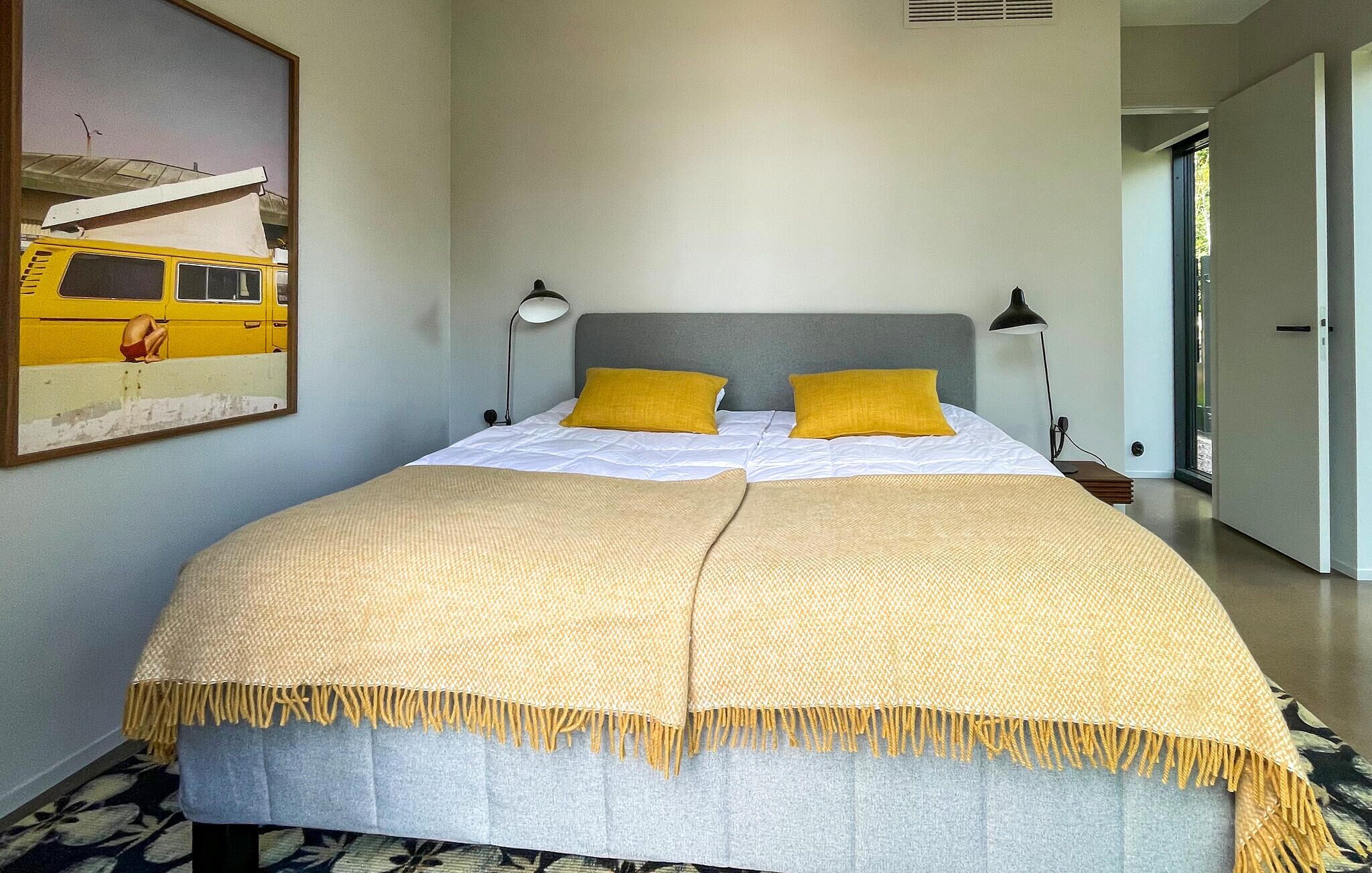 Una cama con cabecera gris, dos almohadas amarillas y una manta color beige con flecos.