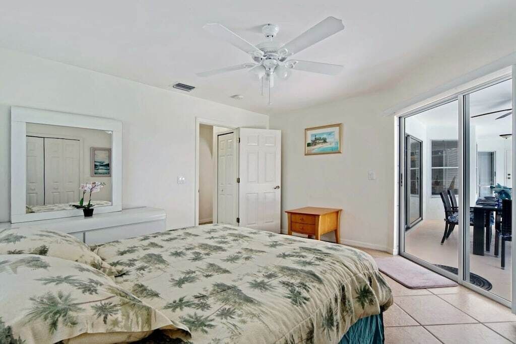 Une chambre à coucher avec un lit, un ventilateur de plafond, une porte, une petite table et une vue sur l’extérieur, visible à travers une porte vitrée.