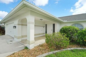 A single-story house with a covered garage, a small porch with columns, and a well-maintained lawn. - CCVS - Villa Allegra (Cape Coral)