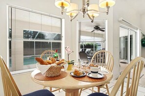 A dining area with a round table, chairs, a chandelier, and a window with blinds. - CCVS - Villa Allegra (Cape Coral)