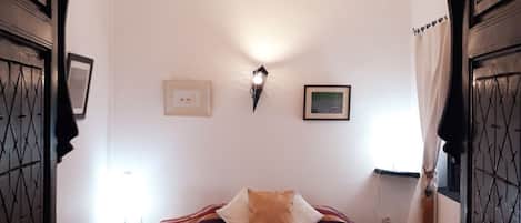 A room with a bed, a chandelier, framed pictures on the wall, and a window with curtains.