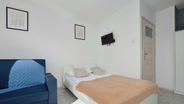A bedroom with a bed, a sofa, a TV, and two framed pictures on the wall. - Cozy Apartment with Bedroom | Smart TV and Washing Machine (Łódź)