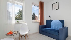 A modern living room with a blue sofa, a white chair, a small table with a bowl of fruit, and two windows with curtains. - Cozy Apartment with Bedroom | Smart TV and Washing Machine (Łódź)