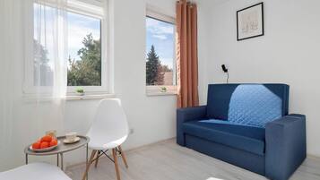 Una sala de estar moderna con un sofá azul, una silla blanca, una mesita con un cuenco de fruta y dos ventanas con cortinas.