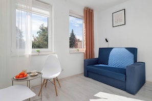 Ett modernt vardagsrum med en blå soffa, en vit fåtölj, ett litet bord med en skål med frukt och två fönster med gardiner.