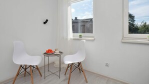 Una habitación pequeña con dos sillas blancas, una mesa con un bol de naranjas y una ventana que ofrece vistas a edificios y árboles.
