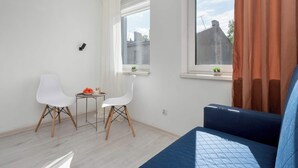 Una habitación pequeña y luminosa con una cama, dos sillas blancas, una mesita y una ventana con cortinas.
