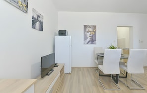Een moderne, minimalistisch ingerichte kamer met een flatscreen televisie, een witte koelkast, een eettafel met witte stoelen en twee kunstwerken in lijsten aan de muur.