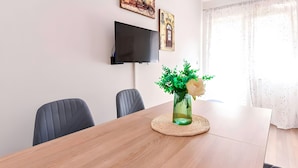 A dining area with a wooden table, chairs, a vase with green foliage and a white flower, and a flat-screen TV mounted on the wall.