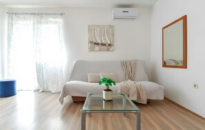 A living room with a sofa, a glass coffee table with a plant, a painting on the wall, and an air conditioning unit.