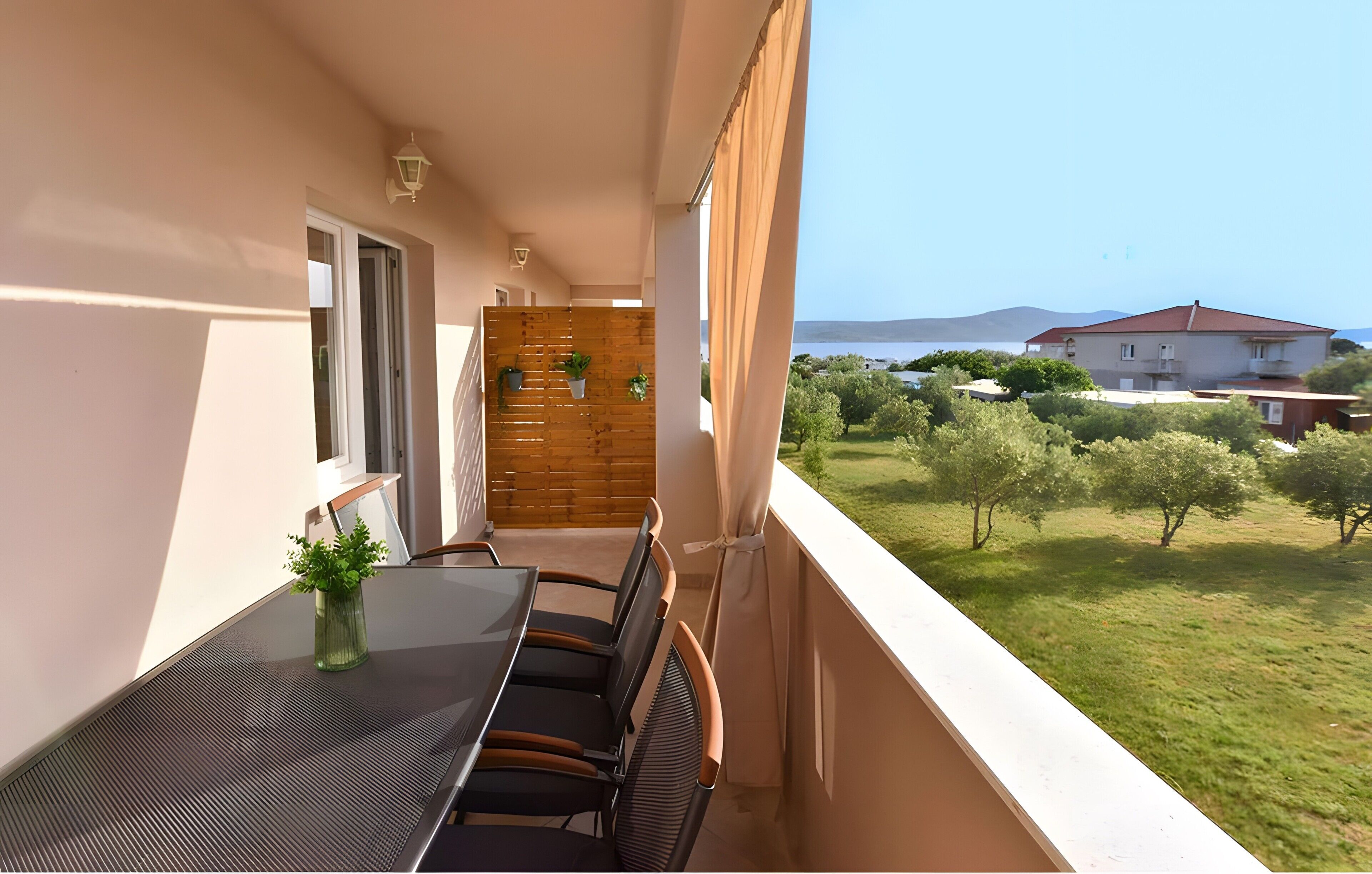 A balcony with a table and chairs, a wooden partition, and a view of a green landscape and buildings in the distance.