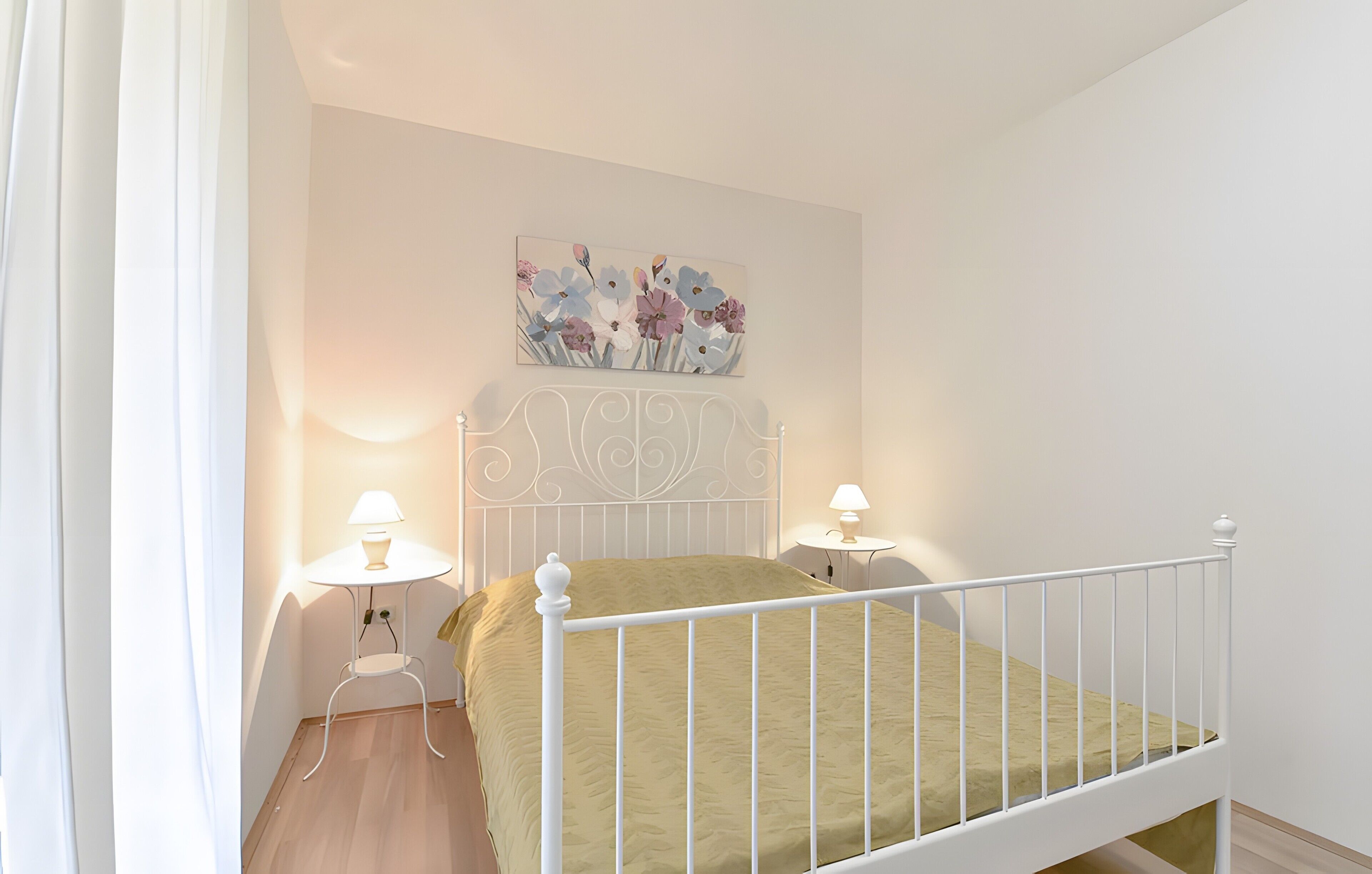 A bedroom with a white metal bed frame, a green bedspread, two bedside lamps, and a floral wall art piece above the bed.