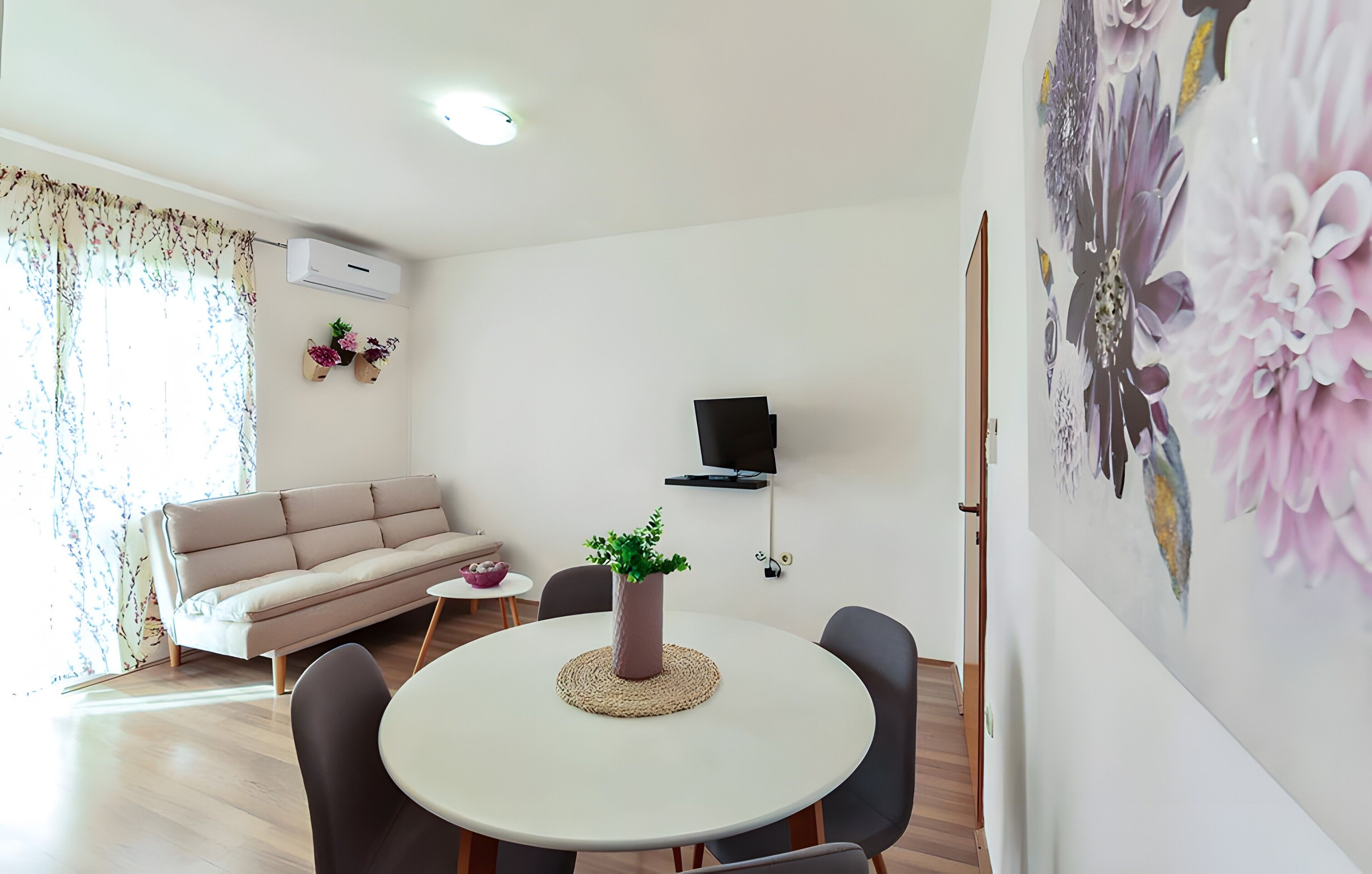 A living room with a sofa, a round dining table with chairs, a potted plant, a flat-screen TV mounted on the wall, and a floral artwork on the wall.