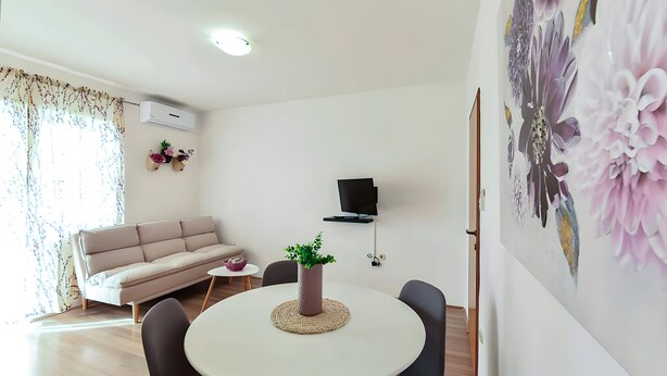 Een woonkamer met een bank, een ronde eettafel met stoelen, een potplant, een flatscreen-televisie aan de muur en een bloemistisch kunstwerk.