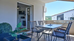 Uma área de refeições ao ar livre coberta, com uma mesa, cadeiras, um vaso de planta e uma casa ao fundo.