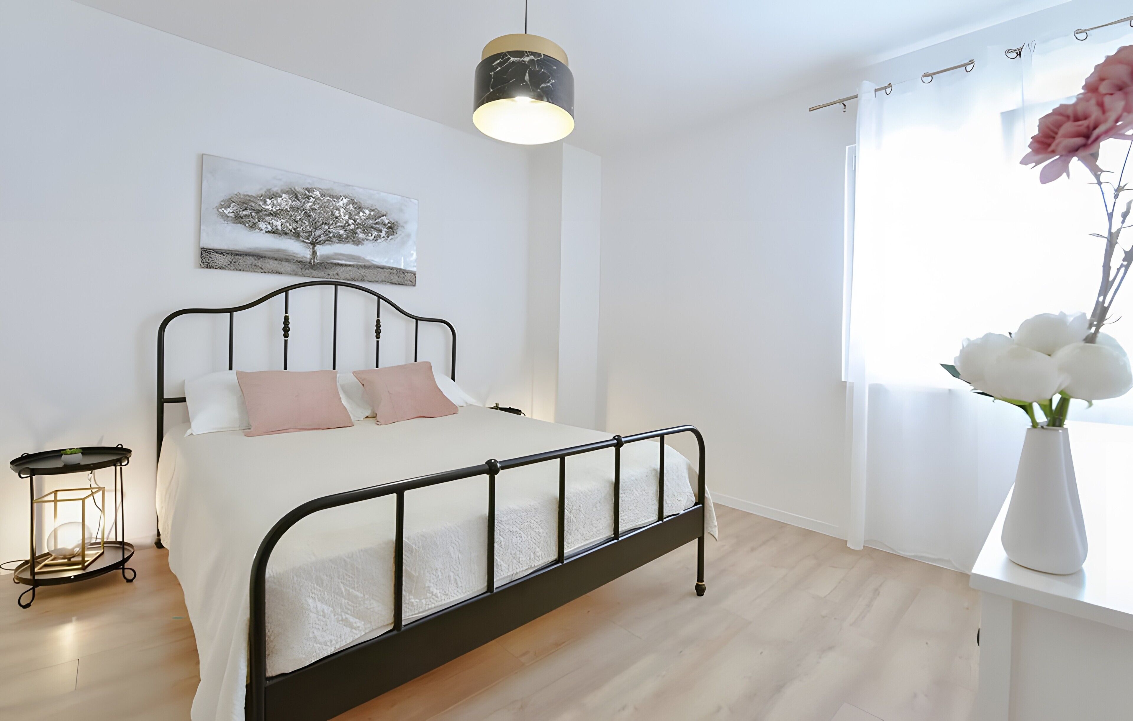 A bedroom with a bed, a bedside table, a hanging light, a window with curtains, and a wall-mounted picture.