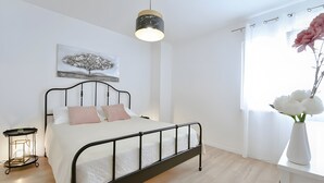 Quarto com cama, mesa de cabeceira, luminária suspensa, janela com cortinas e um quadro na parede.