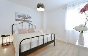 Ein Schlafzimmer mit Bett, Nachttisch, Pendelleuchte, einem Fenster mit Vorhängen und einem Wandbild.