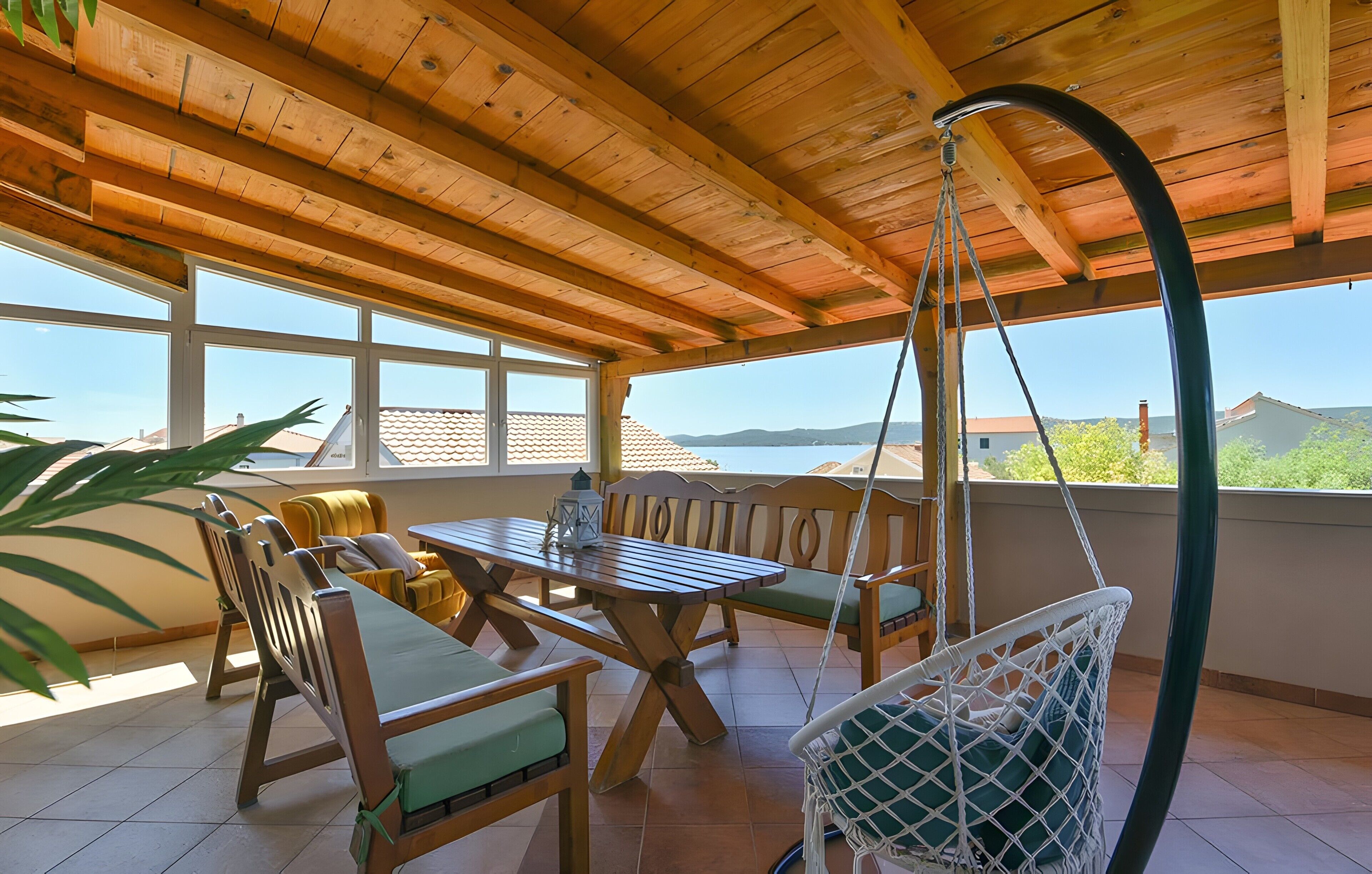 A covered outdoor patio with a wooden table and chairs, a hanging swing, and a view of the sea.