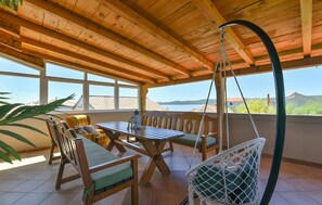 Een overdekt terras buiten met een houten tafel en stoelen, een hangschommel en uitzicht op zee.