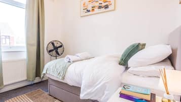 Hotellihuone, jossa on sänky, yöpöytä kirjoineen ja pullo, tuuletin ja ikkuna, jossa on verhot.