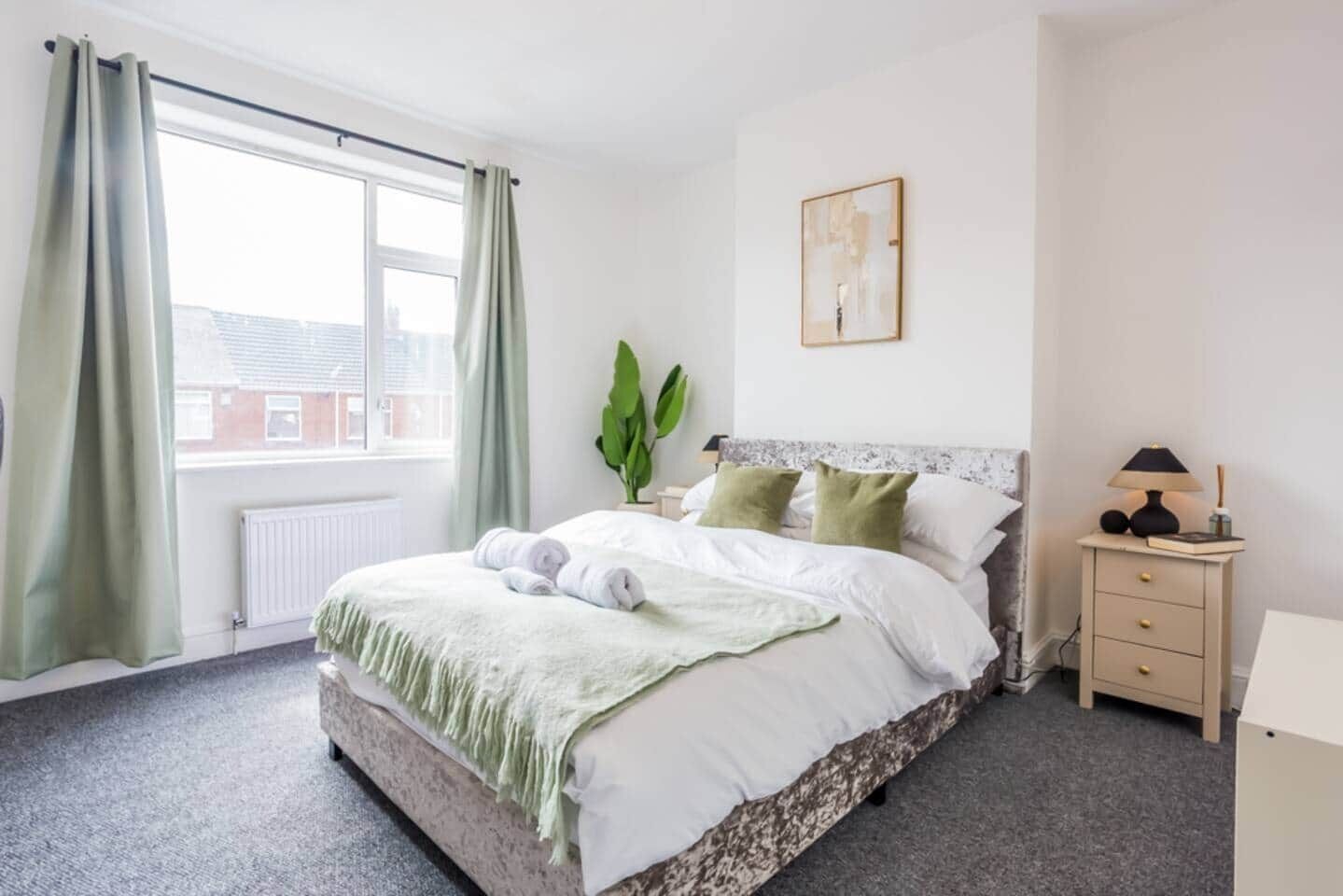 Un dormitorio con cama, mesita de noche, lámpara, planta y ventana con cortinas.