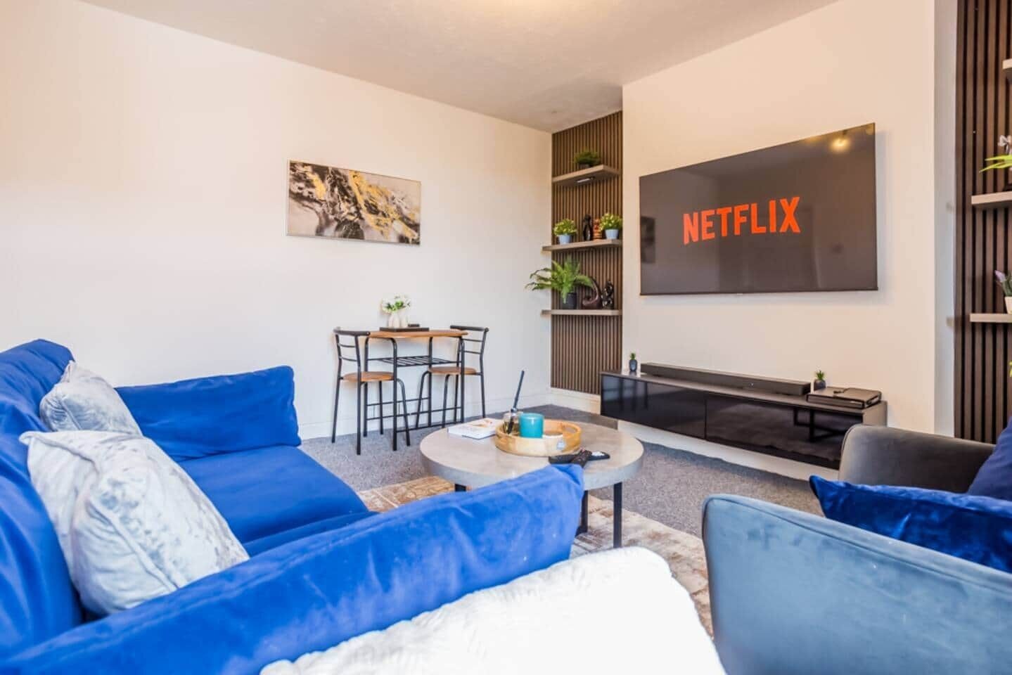 Moderni olohuone, jossa on sininen sohva, sohvapöytä, televisio, jonka näytöllä näkyy teksti ”NETFLIX”, ja pieni ruokailutila.