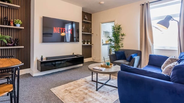 A modern living room with a flat-screen TV mounted on the wall, a sofa, a coffee table, and a dining area with a wooden table and chairs. - The Geordie Getaway! (Newcastle upon Tyne)