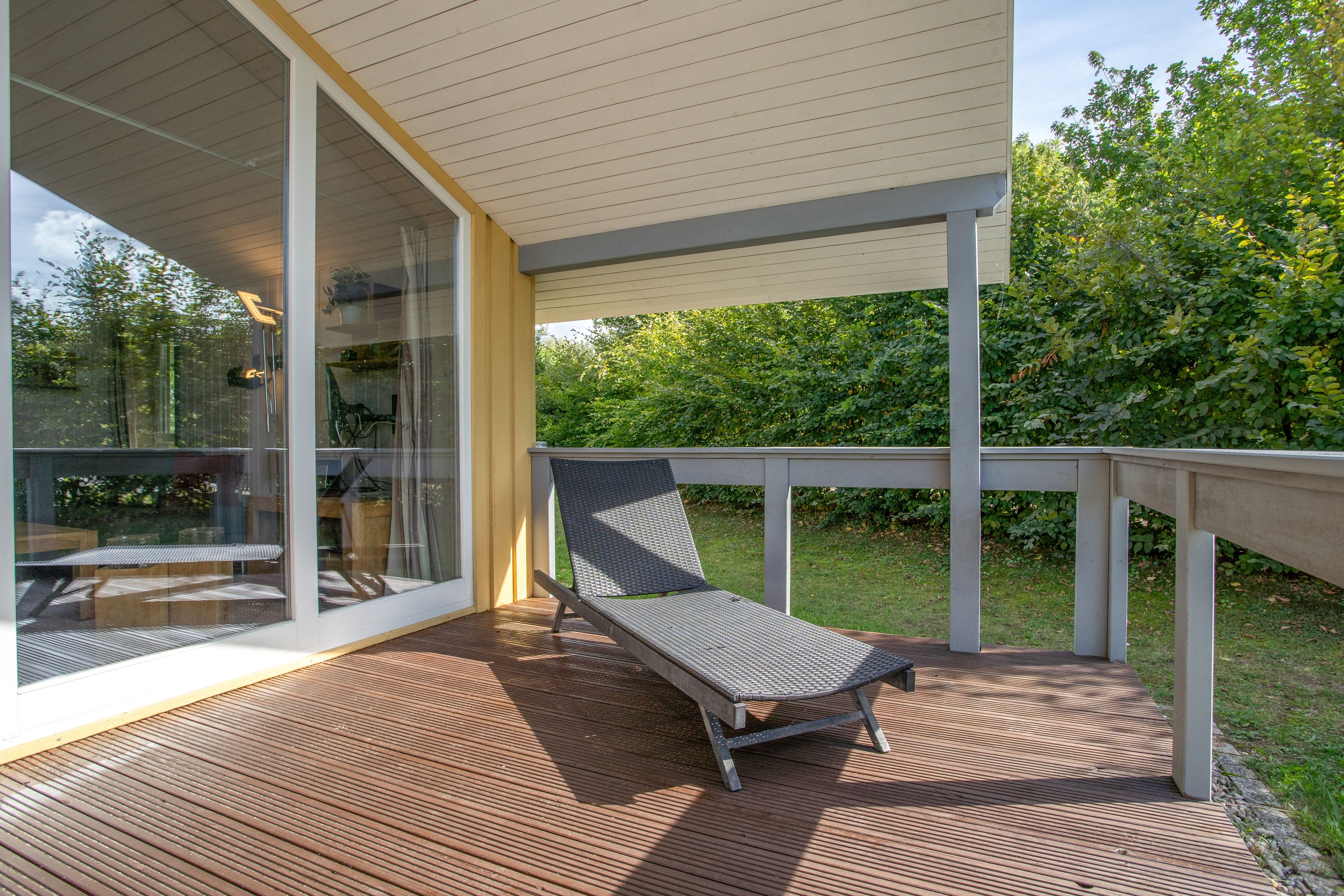 Un espace extérieur abrité, doté d’une terrasse en bois, d’un fauteuil et d’une porte vitrée donnant sur un espace intérieur.