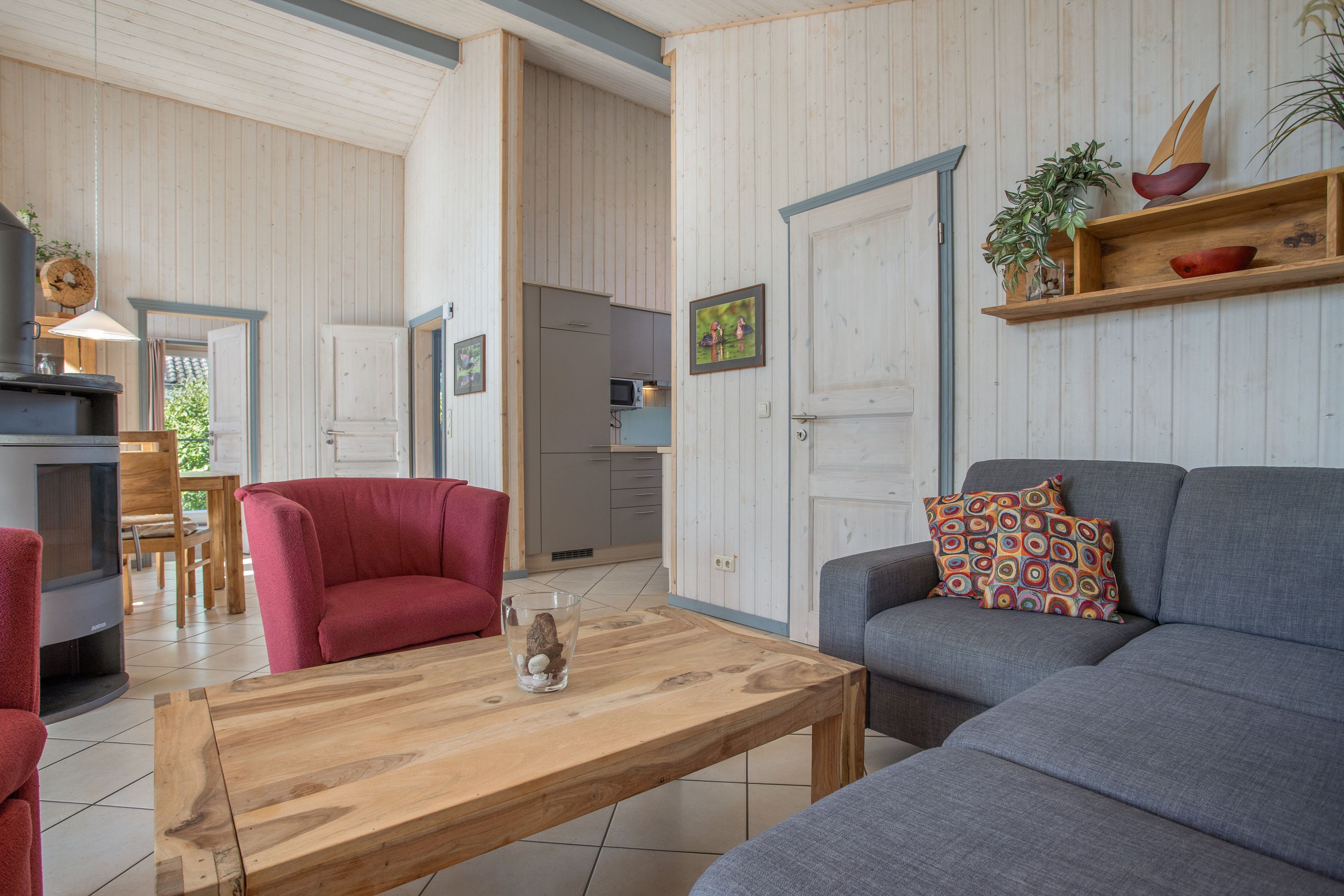 A living room with a sofa, a red armchair, a wooden coffee table, and a kitchen in the background.