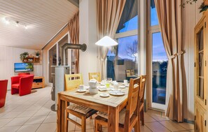 A dining area with a wooden table set for breakfast, a television in the background, and large windows with curtains. - Holiday Home '55 Typ Eisvogel' with Private Terrace, Private Garden and Wi-Fi (Rechlin)