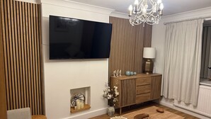 Ein modernes Wohnzimmer mit Flachbildfernseher, Wandpaneelen aus Holz, einer Kandelaberleuchte, einem Holztisch und einer Kommode mit Dekoration.