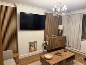 A modern living room with a flat-screen TV, wooden panel wall, a chandelier, a wooden coffee table, and a sideboard with decorative items. - House Sleeps 4 With Parking and Garden (Scunthorpe)