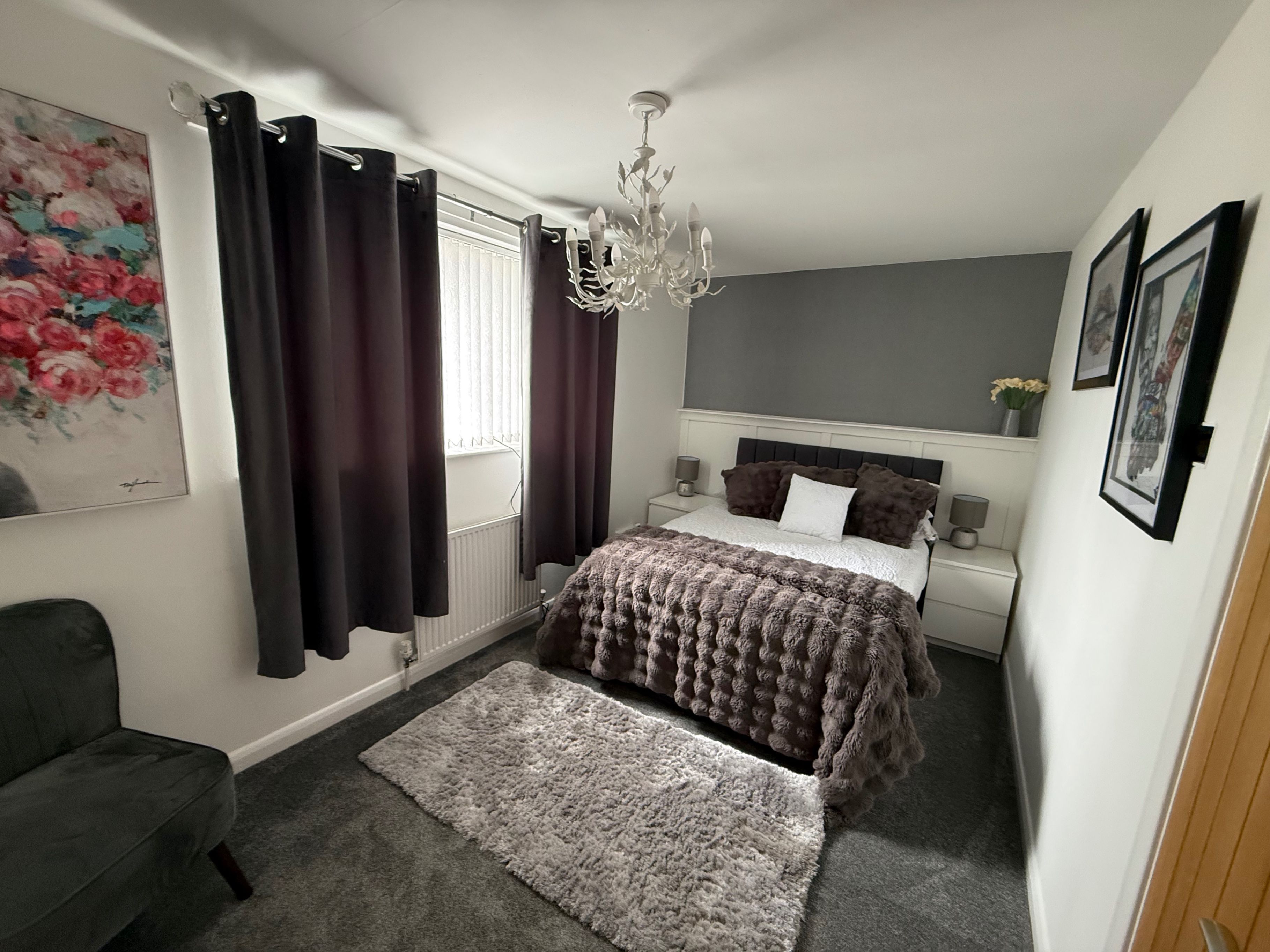 A bedroom with a bed, a chandelier, a grey sofa, a rug, and a painting on the wall.