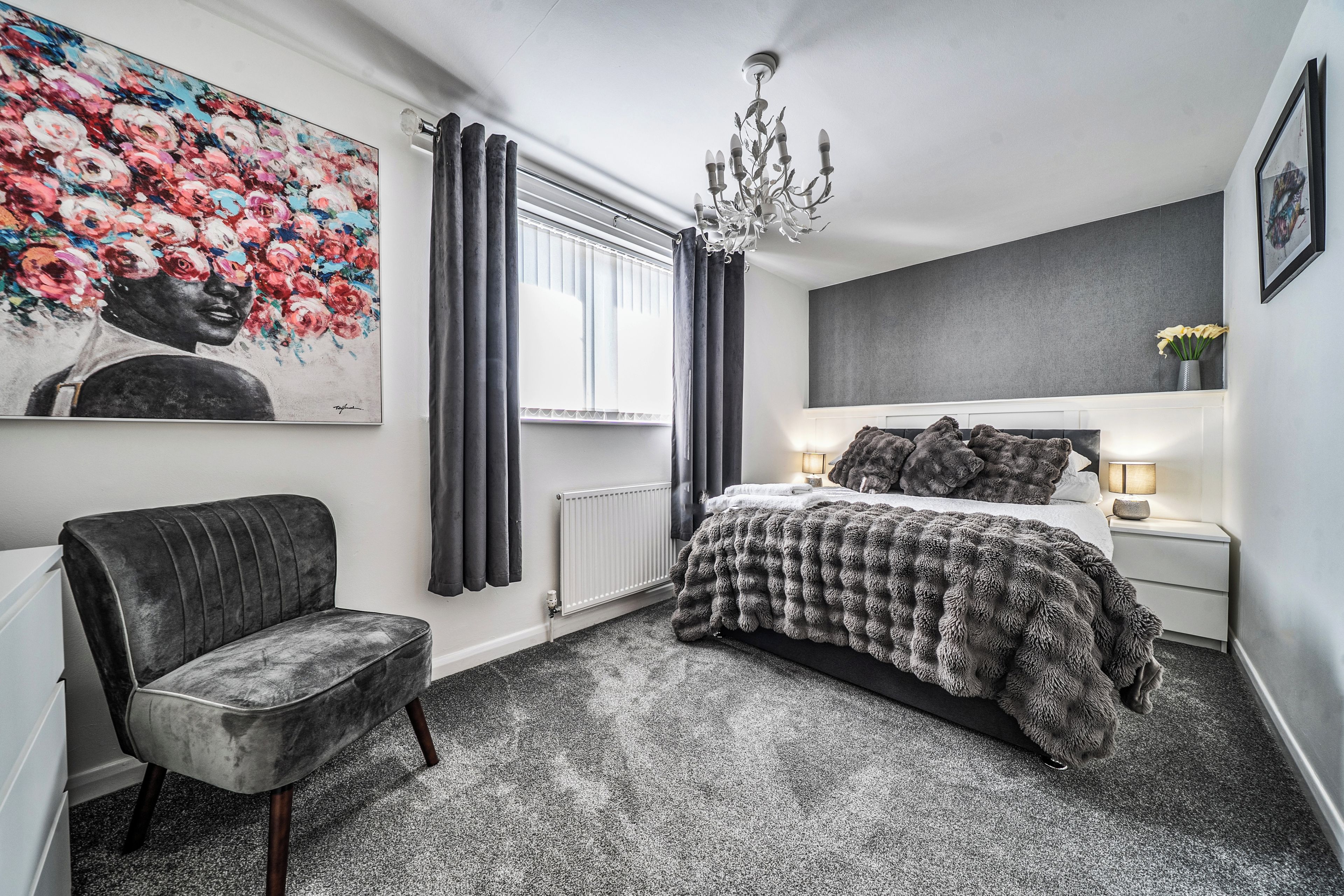 A bedroom with a bed, a chair, a radiator, a window with curtains, a chandelier, and a painting on the wall.