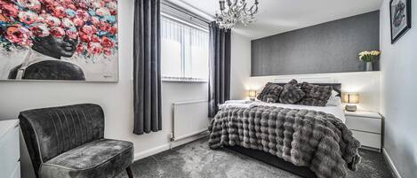 A bedroom with a bed, a chair, a radiator, a window with curtains, a chandelier, and a painting on the wall.