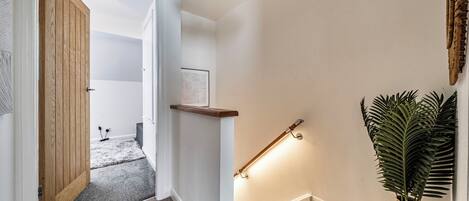 A hallway with a wooden door, a wall-mounted shelf, a framed picture, a floor lamp, and a potted plant.