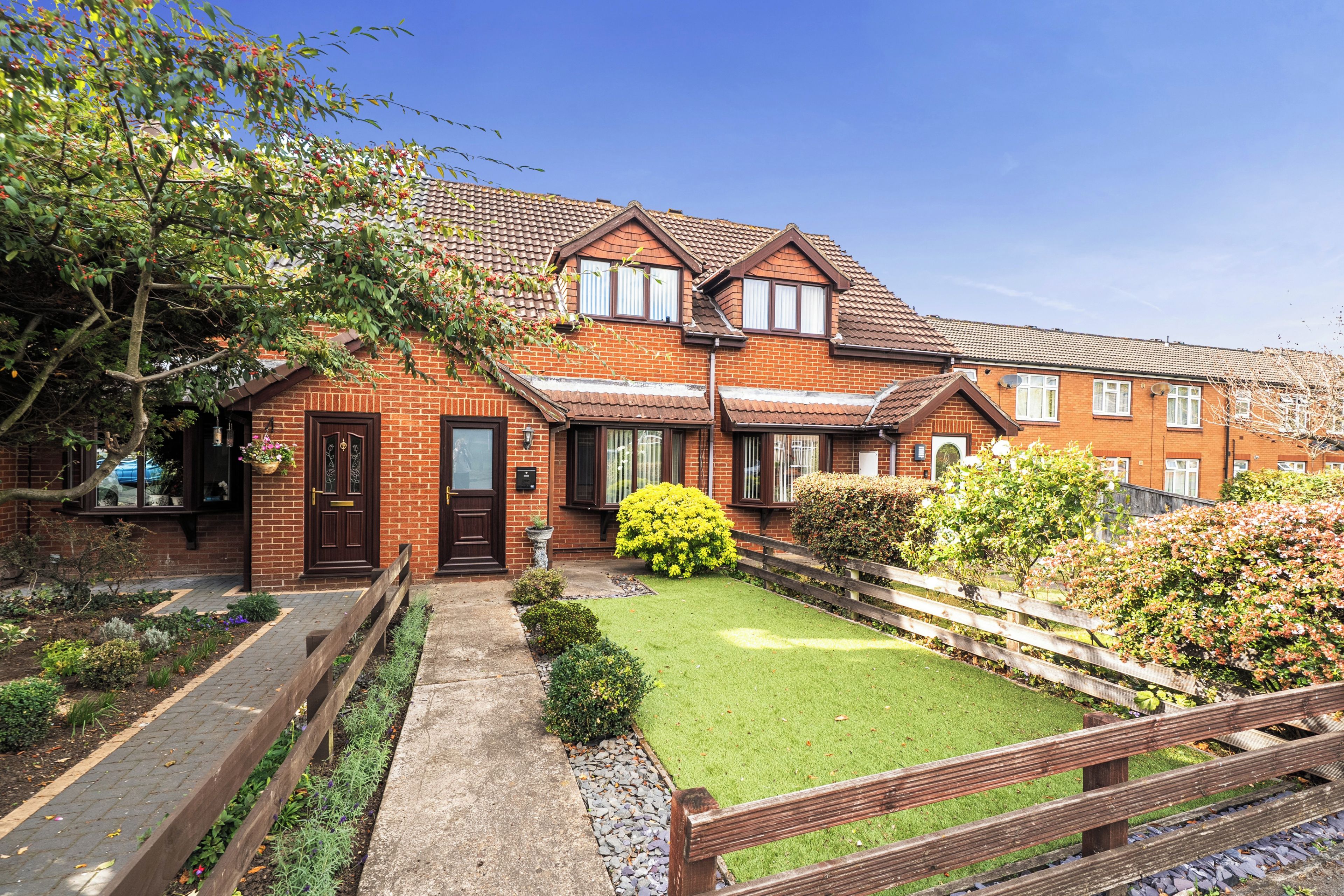 A two-story brick house with a front garden, a wooden fence, and a paved driveway.