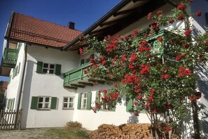 Un bâtiment à deux étages, avec un toit recouvert de tuiles rouges, des volets verts et un balcon orné de fleurs rouges.
