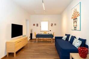 Ein Wohnzimmer mit einem Flachbildfernseher, einem Esstisch mit Stühlen, einem blauen Sofa und einer roten Dekorationsvase.