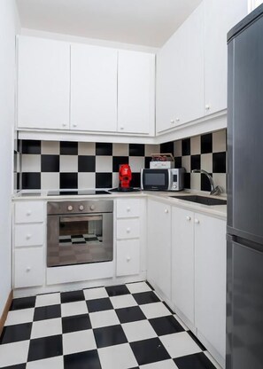 Eine kompakte Küche mit weißen Schränken, einer schwarz-weißen Schachbrett-Spritzschutzleiste, einer Mikrowelle, einer roten Kaffeemaschine und einem Edelstahl-Kühlschrank.