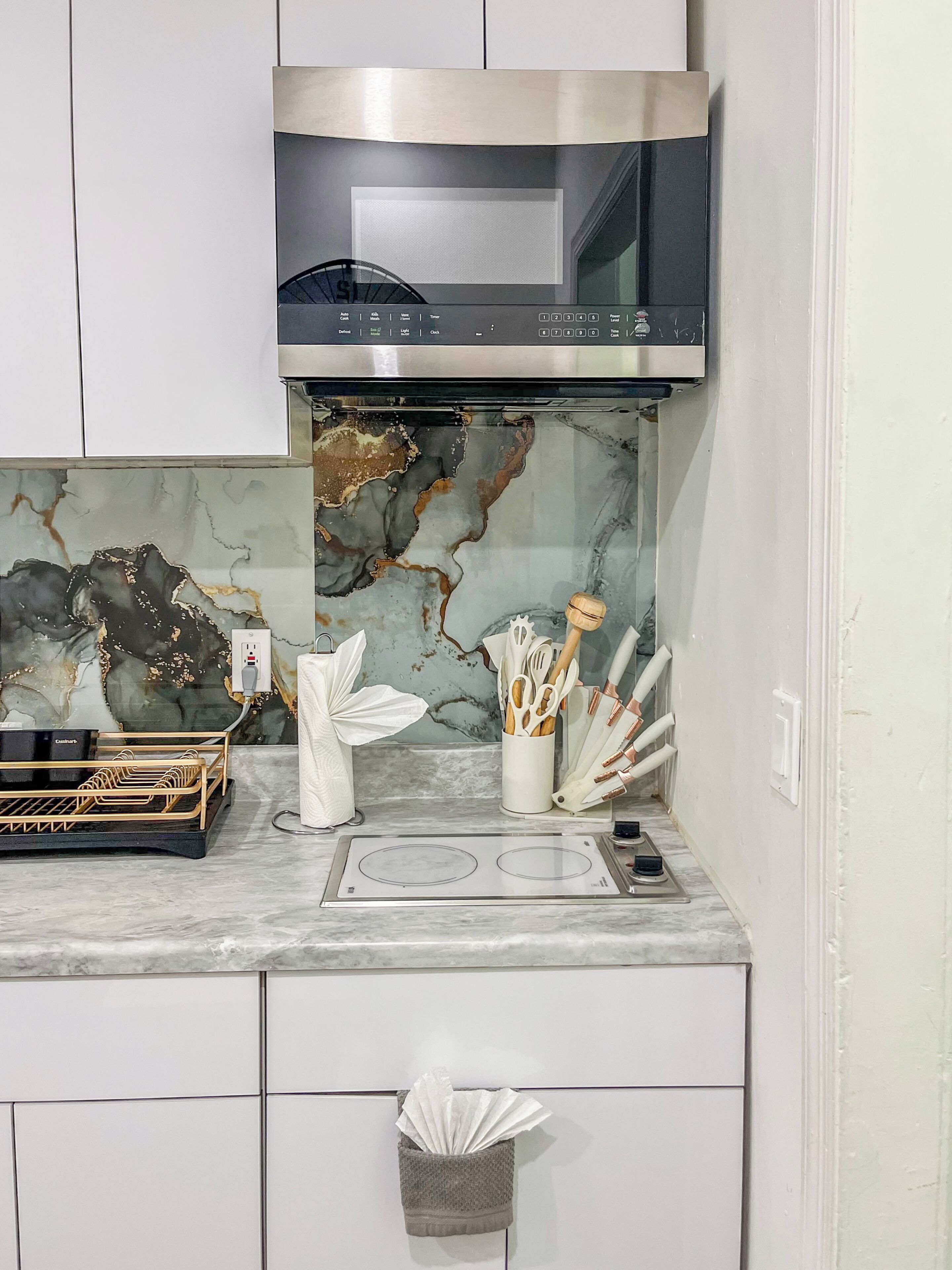 A kitchen with a stainless steel microwave, a marble backsplash, and a countertop with a dish rack and utensils.