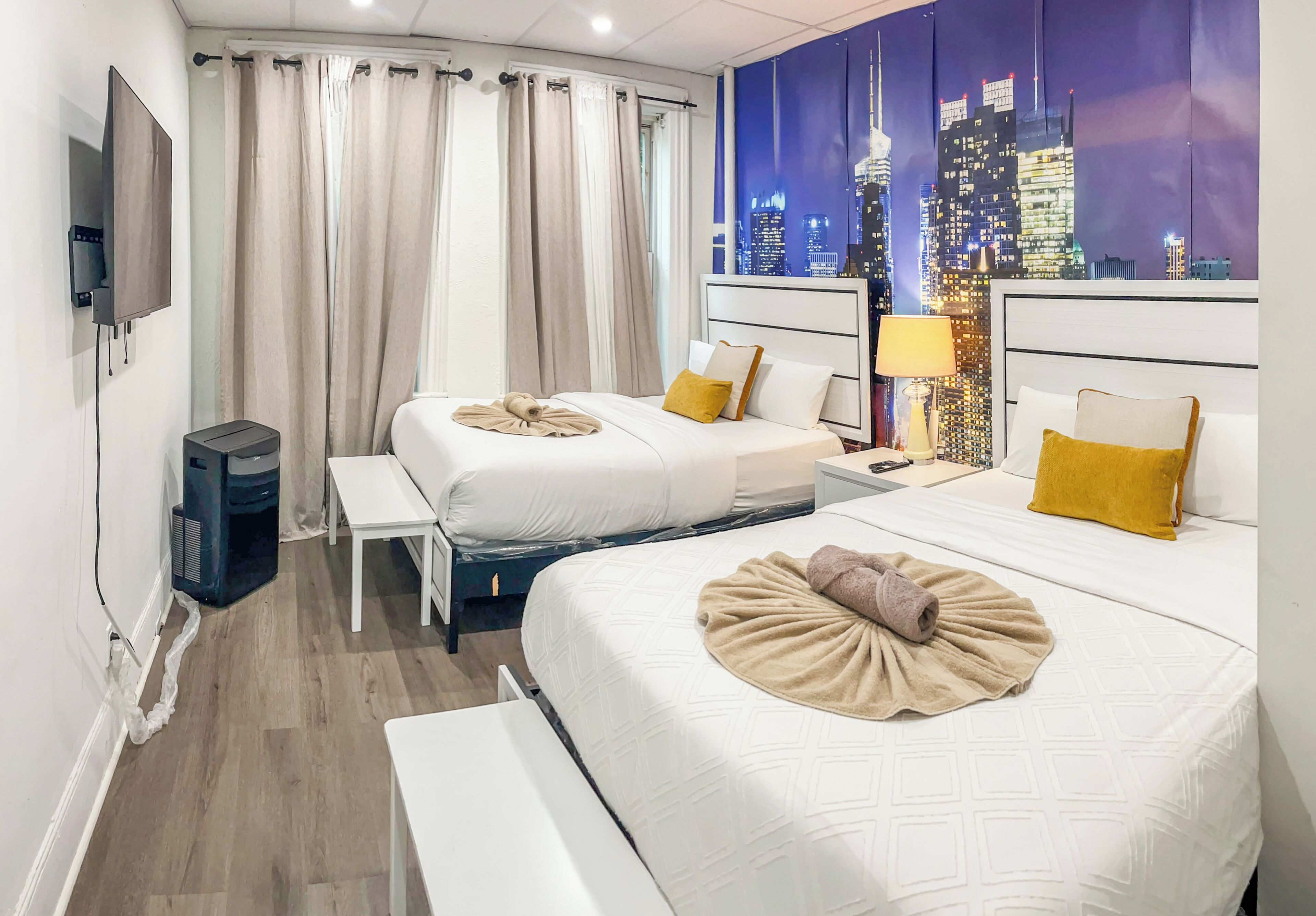 Une chambre d’hôtel avec deux lits, un bureau, une chaise, un téléviseur et une fresque représentant un paysage urbain sur le mur.